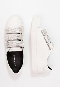 Baskets blanches avec une tige en cuir, trois rangées de brides décoratives ornées de strass et une semelle en caoutchouc texturée. La doublure intérieure est noire.