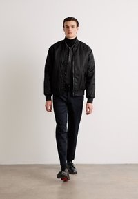 Svart bomberjakke med glidelås foran, båret over en tettsittende svart turtleneck, kombinert med skreddersydde marineblå bukser og svarte sko. Enkel design.