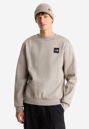 Ung mand iført beige strikket hue, lysegrå The North Face sweatshirt og sorte cargobukser, står med den ene hånd i lommen.