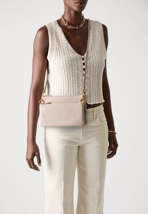 Borsa a tracolla in pelle beige con una finitura testurizzata, hardware dorato e una tracolla rimovibile, abbinata a un top in maglia crema e pantaloni chiari.