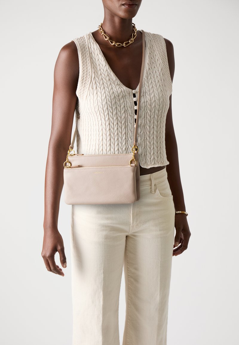 Beige leather crossbody bag with a textured finish, gold hardware, and a detachable strap, paired with a cream knit top and light pants.