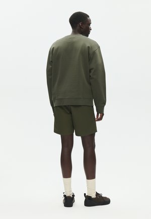 Mies, jolla on oliivinvihreä pitkähihainen collegepaita ja siihen sopivat shortsit, valkoiset sukat ja tummat lenkkarit, seisoo selin valkoista taustaa vasten.