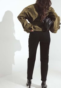 Chaqueta de ante color verde oliva, corta y con mangas anchas, llevada sobre unos pantalones vaqueros negros de talle alto y botines negros con tacón. Textura suave y diseño minimalista.