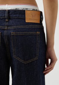 Jeans de denim azul oscuro con una etiqueta de cuero en la parte trasera, costuras doradas y dos bolsillos traseros. La cinturilla presenta un forro con un patrón.