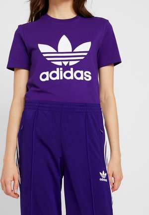 Femme portant un t-shirt Adidas violet et un pantalon assorti violet avec des logos et des bandes Adidas blanches, debout devant un fond uni.