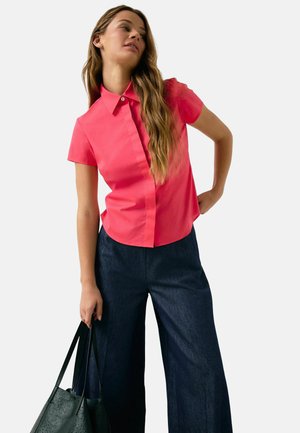Short-sleeve, collared shirt in bright pink, paired with wide-leg dark denim pants. Model holding a textured black tote bag.