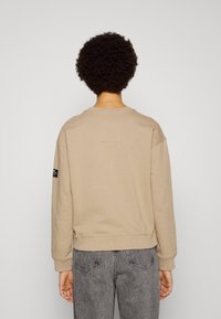 Sudadera beige con cuello redondo, puños y dobladillo acanalados. Presenta una pequeña etiqueta en la manga izquierda. Combinada con pantalones grises.