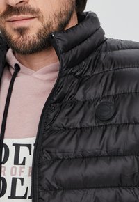 Chaleco acolchado negro con collar alto y cierre de cremallera, que presenta una insignia de logo circular en el pecho y que se lleva sobre una sudadera rosa.