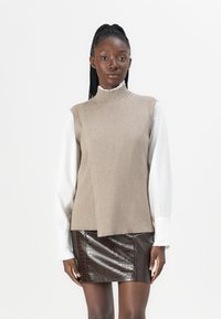 Pull en laine côtelé beige sans manches avec un col roulé par-dessus une chemise blanche à volants à manches longues, associé à une mini-jupe en cuir crocodile marron.
