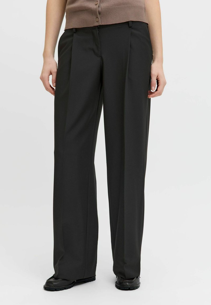 Asmuo, dėvintis laisvus juodus kelnes ir juodus batus, stovintis prieš balto fono sieną, rankos laisvai nuleistos šonu.