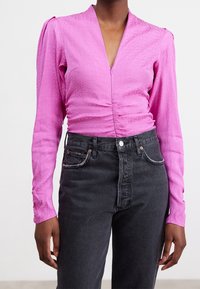 Persona che indossa un top rosa fitted a maniche lunghe con un motivo sottile e jeans scuri a vita alta, in piedi davanti a uno sfondo semplice.