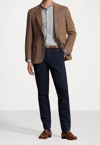 Uomo in giacca herringbone marrone, camicia button-up grigio chiaro, pantaloni blu navy, cintura in pelle marrone e mocassini, e orologio da polso, in piedi.