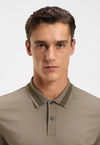 Oliven grønn polo skjorte i glatt stoff med ribbestrikket krage, tre-knapp placket og en liten logo på venstre side.
