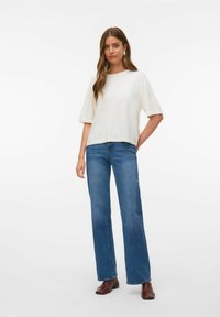 Lysecremet kortærmet top med rund halsudskæring, parret med højtaljede blå brede jeans. Modellen har brune firkantede sko på.