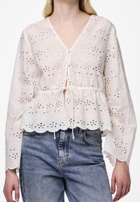 Blouse blanche brodée avec un col en V, des manches bouffantes et une taille à cordon ; présente un ourlet à volants et des motifs floraux découpés.