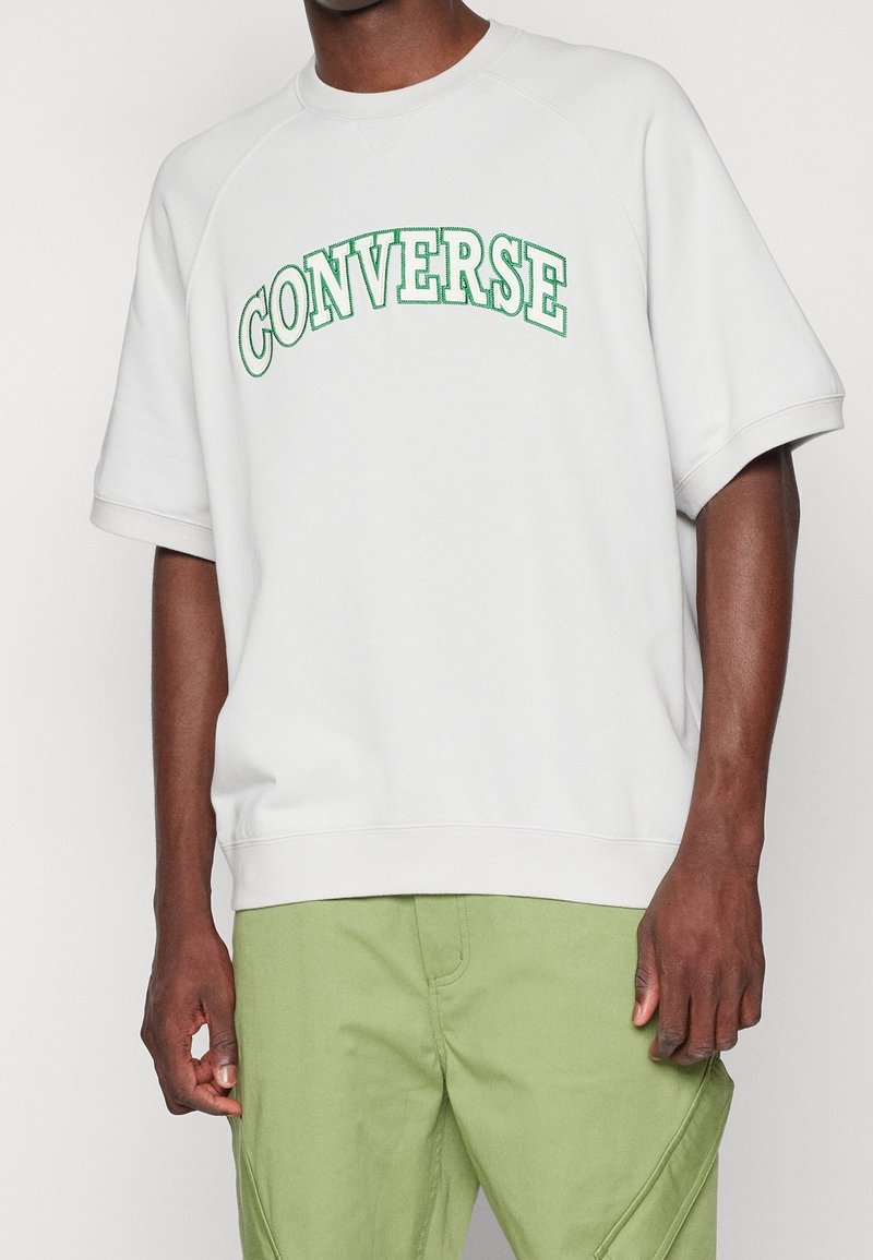 Biała bluza z krótkim rękawem, z logo "CONVERSE" w zielonym łukowym nadruku oraz luźnym krojem. Miękka faktura z ściągaczami przy rękawach i u dołu.