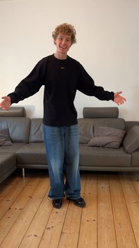 Black sweatshirt, blue wide-leg jeans, and dark shoes. Standing with arms outstretched in a room with a gray sofa and wooden floor.