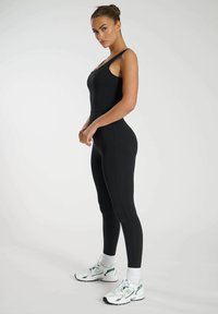 Zwarte aansluitende bodysuit met brede bandjes, naadloos ontwerp en een hoge taille. Gepaald met witte sportschoenen met groene accenten.