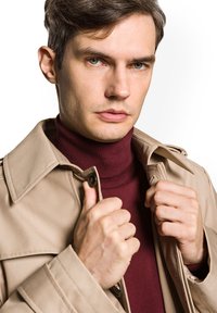 Beżowy trencz z podwójnymi guzikami na tle bordowego golfu. Gładki materiał z usztywnionym kołnierzem i widocznymi detalami przeszycia.