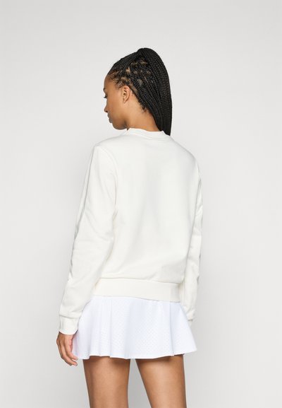 Un sweat-shirt blanc à col ras du cou avec des manches longues, une texture lisse et des poignets côtelés, porté avec une jupe blanche à superposition perforée.