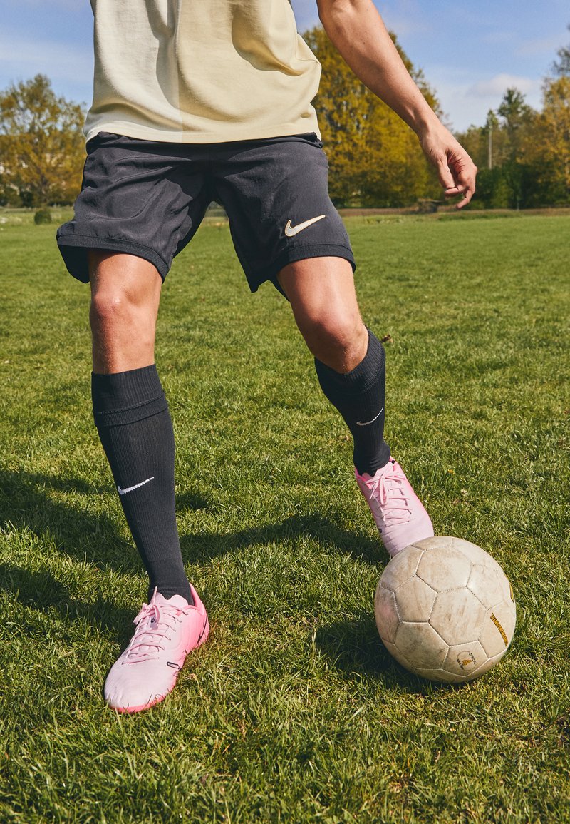 Shorts Nike noirs avec logo doré, chaussettes noires jusqu'aux genoux et chaussures de football roses, positionnés à côté d'un ballon de football usé sur l'herbe.
