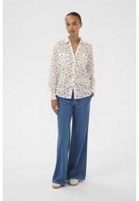 Blouse légère et transparente à motif floral, avec un devant boutonné et deux poches poitrine. Portée avec un jean en denim bleu à jambes larges.
