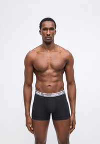 Homme musclé debout devant un fond uni, portant un boxer Nike noir avec une ceinture grise et un logo visible de face, les bras détendus.
