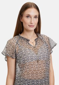 Transparante blouse in bruin en wit met een patroon, met gerimpelde mouwen, een keyhole-halslijn en een gouden accentdetail bij de halslijn.