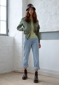 Zelen bomber jakna, svetlo zelena majica, svetlo modri zvitki jeans, črne gležnjarje in črna klobuček, na ozadju bele stene.