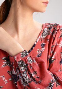 Rød blomstret bluse med V-udskæring, puffede ærmer og knapdetalje ved manchetten, lavet i et tekstureret stof med indviklede blomstermønstre.