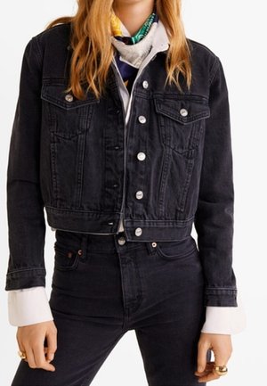 Person iført en sort cropped denimjakke over en hvid skjorte og sort jeans med høj talje, accessoriseret med ringe og et farverigt tørklæde.