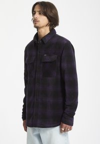 Camisa de franela a cuadros en negro y morado con cierre de botones en el frente, dos bolsillos en el pecho y una textura suave. Presenta un cuello y mangas largas.