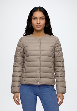 Femme aux longs cheveux foncés portant une veste matelassée marron clair avec des boutons-pression et un jean bleu, debout devant un fond blanc uni.