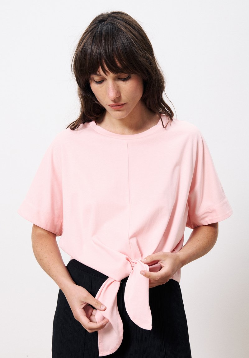 T-shirt en coton rose clair avec des manches courtes, un col rond et un détail noué à la taille, associé à un pantalon noir texturé.