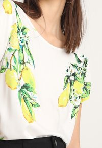 Camicetta bianca a maniche corte con una vivace stampa di limoni e fiori in verde e giallo. Tessuto morbido con scollatura rotonda e vestibilità comoda.