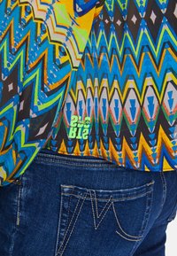 Short-sleeve shirt with a vibrant geometric pattern in blue, yellow, and green, layered over dark denim jeans with detailed stitching.
