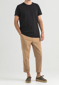 Schwarzes Baumwoll-T-Shirt mit kurzen Ärmeln, kombiniert mit khakifarbenen, verkürzten Hosen. Strukturiertes, dunkelbraunes Schuhwerk, Schnürsenkel gebunden, sichtbares Knöchel.