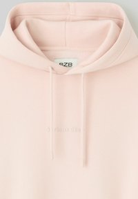 Haut à capuche rose clair avec une texture douce, doté d'une capuche à cordon de serrage et d'un logo brodé "Serious Club" sur le devant.