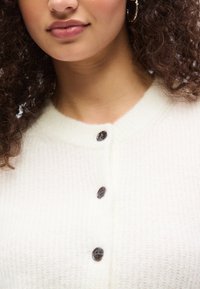 Γυναίκα που φοράει λευκό πλεκτό ζακέτα με μαύρα κουμπιά, με χρυσό κρίκο στο αυτί ορατό, και σγουρά μαλλιά που πλαισιώνουν το πρόσωπό της.