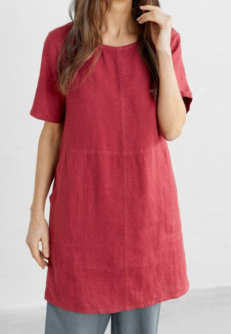 Femme portant une tunique rouge à manches courtes et ample par-dessus un pantalon gris, debout devant un fond clair uni.