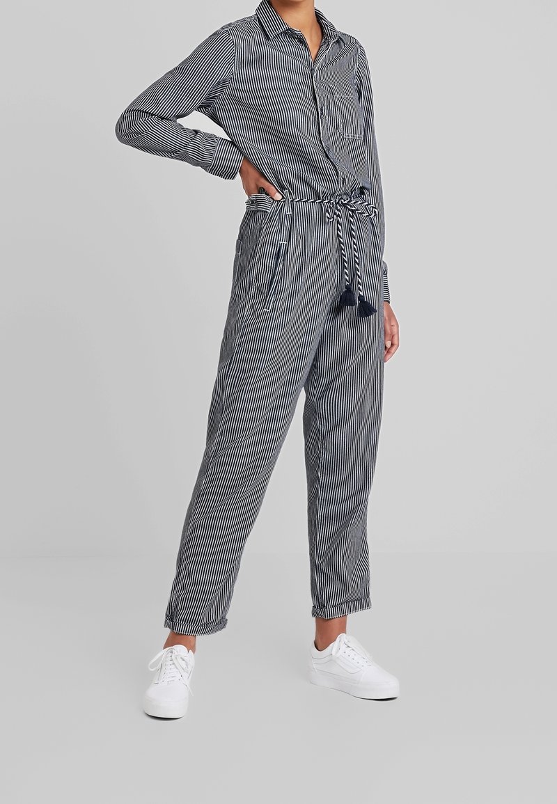 Randig jumpsuit med knapp-up överdel, långa ärmar och knytmidja. Har två sidofickor och en bröstficka, tillverkad av lättviktig tyg.