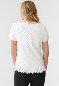 Top alb cu un design texturat, plisat, având un efect de valuri, cu mâneci scurte și o margine ondulată. Purtați-l cu pantaloni negri.
