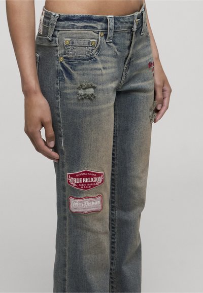 Personne portant un jean bleu délavé avec usures et deux patchs rouge et blanc de la marque True Religion sur la cuisse.