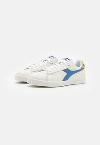 Vita lädersneakers med blå sidostreck och orange detaljer på hälarna, vita snören och logotyp på tungan mot en vit bakgrund.