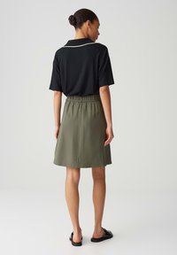 Woman standing facing away wearing a black short-sleeve top with white trim, an olive green knee-length skirt, and black slip-on shoes.