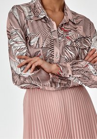 Blusa a maniche lunghe rosa con motivi a foglia neri e bianchi e righe orizzontali, con due tasche davanti e chiusura con bottoni. Abbinare a una gonna plissettata.