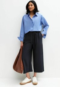 Ljusblå knappad skjorta med en ficka; svarta cropped byxor med dragsko; brun vävd tote-väska; vita sneakers med bruna detaljer.