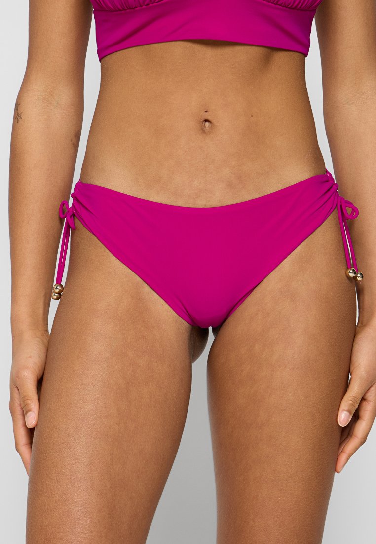 Fuksia bikinitasku, valmistettu sileästä kankaasta, jossa on sivunauhat ja kultaiset helmiakennot. Ominaisuuksiin kuuluu matala vyötäröleikkaus ja minimaalinen peitto.