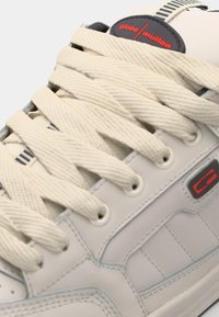Gray leather sneaker featuring beige laces, black and red accents, and a textured design with stitching and eyelet details.