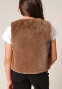 Gilet en fausse fourrure marron avec une texture douce, bords arrondis et sans quincaillerie visible, porté sur un t-shirt blanc, associé à un pantalon noir.